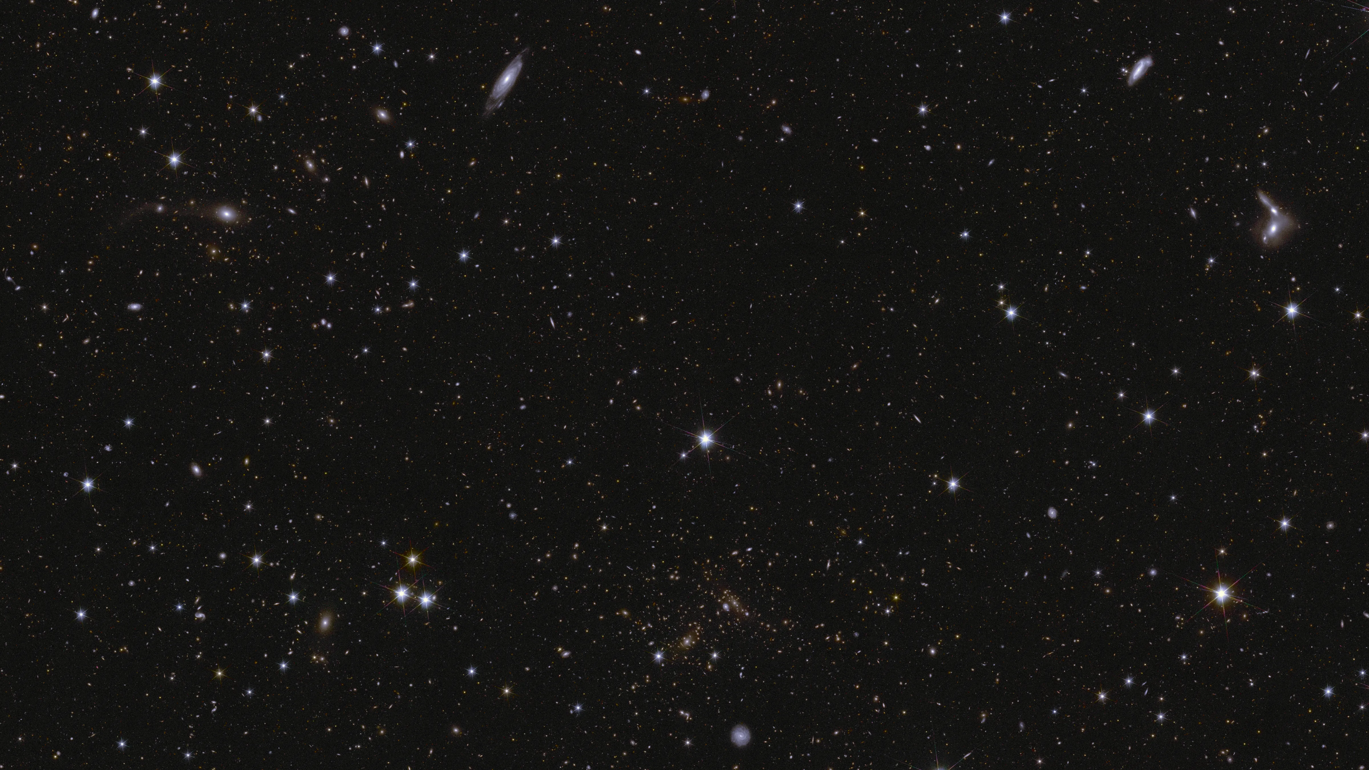 Ein Meer aus Sternen und Galaxien funkelt vor einem schwarzen Hintergrund. Mehrere Galaxien lassen sich an ihrer länglichen Form und/oder ihren Spiralarmen erkennen. Einige Galaxien sind von der Seite zu sehen, während eine markante Spiralgalaxie im unteren Zentrum frontal zu sehen ist. Ganz rechts, zwischen der Mitte und dem oberen Teil des Bildes, befinden sich einige interagierende Galaxien. Auch Galaxienhaufen sind zu sehen, insbesondere in der Nähe des unteren Zentrums, wo verschmierte Strukturen in Bögen auf Gravitationslinsenwirkung hinweisen. Die hellsten Sterne im Bild weisen Beugungsstrahlen auf.