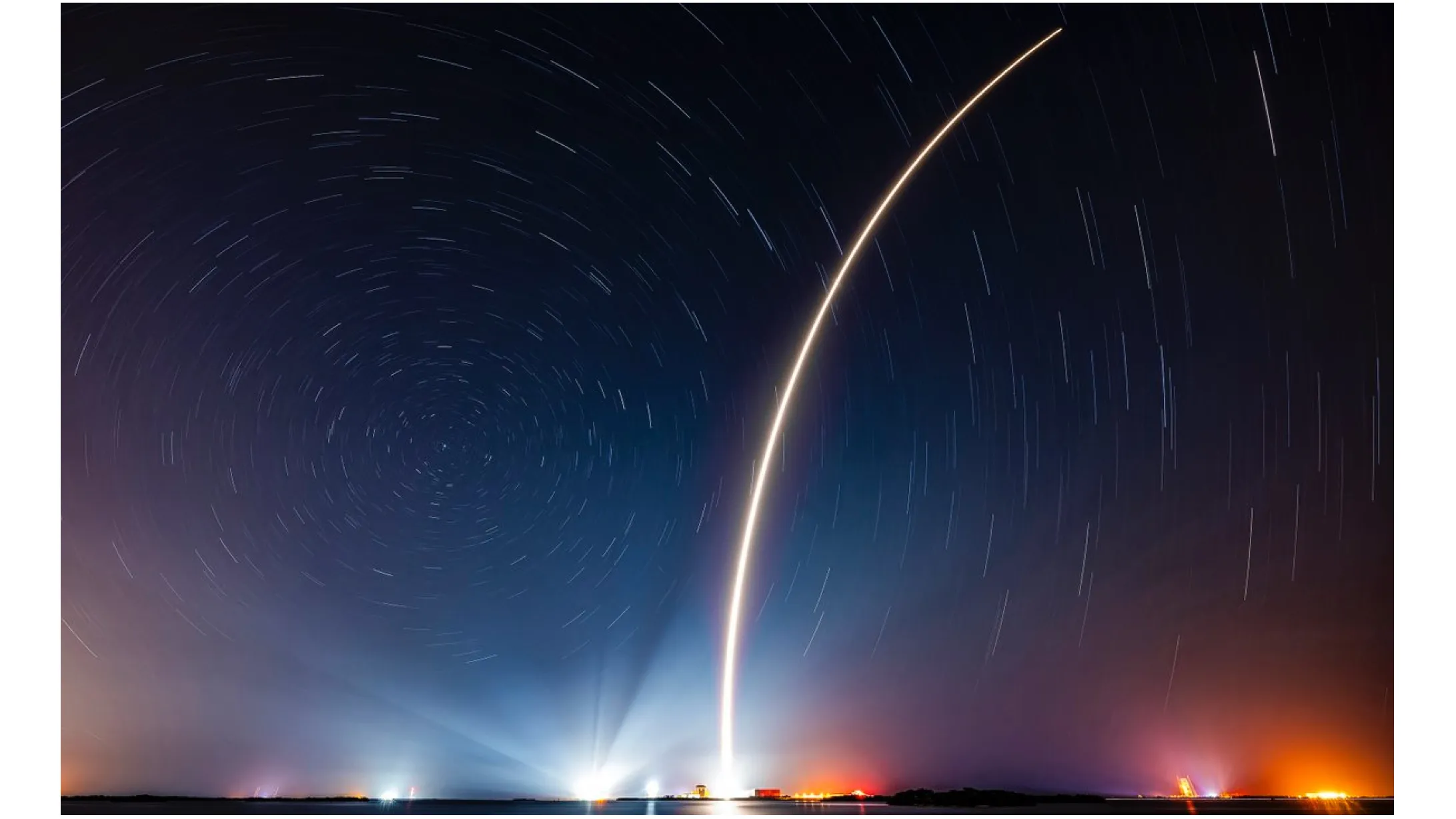Ein Raketenstart zeichnet einen orangefarbenen Bogen in den dunklen Nachthimmel auf diesem Langzeitbelichtungsfoto.