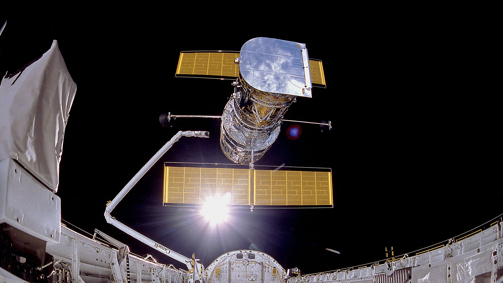 ein großer, silberner Satellit mit orangefarbenen Solarzellenflügeln wird von einem Roboterarm in das grelle Sonnenlicht entlassen